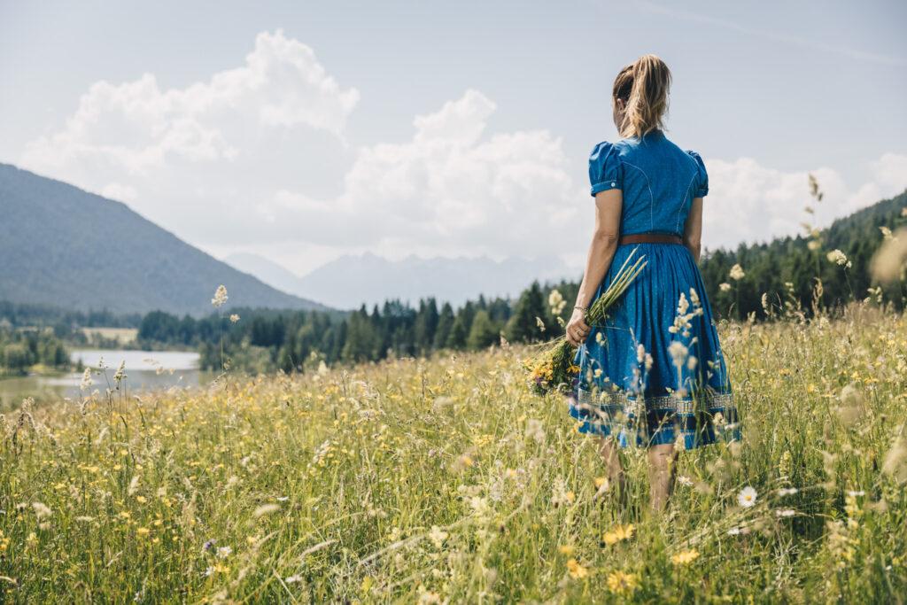 A woman in a blue dress stands on a sunlit meadow in the Seefeld region of Austria, holding wildflowers in her hands. The Alps and clouds can be seen in the background, creating a calm, peaceful atmosphere.