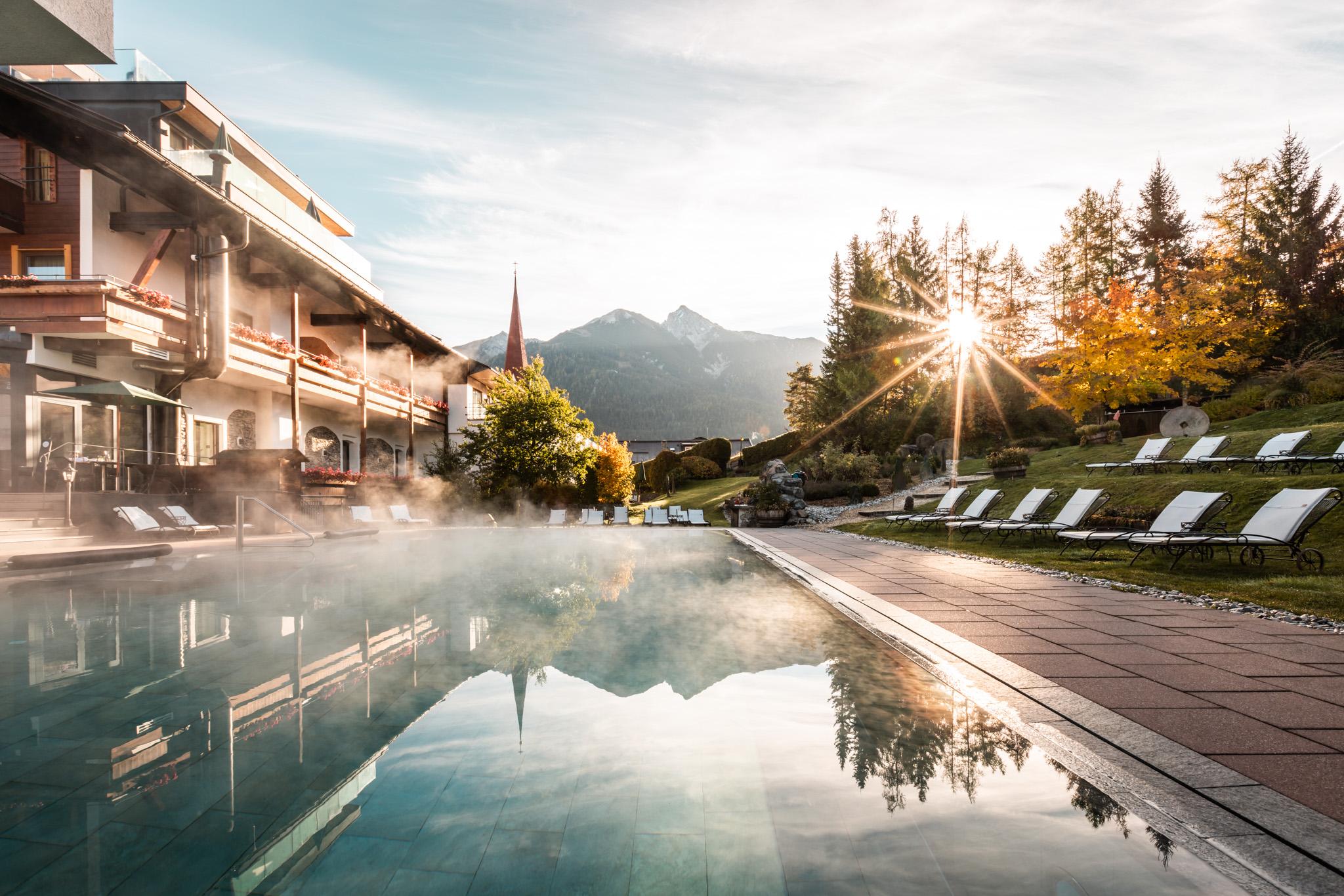 The outdoor pool in autumn. The hotel can still be seen on the left, with deck chairs on the right. The sun shines low through the trees.