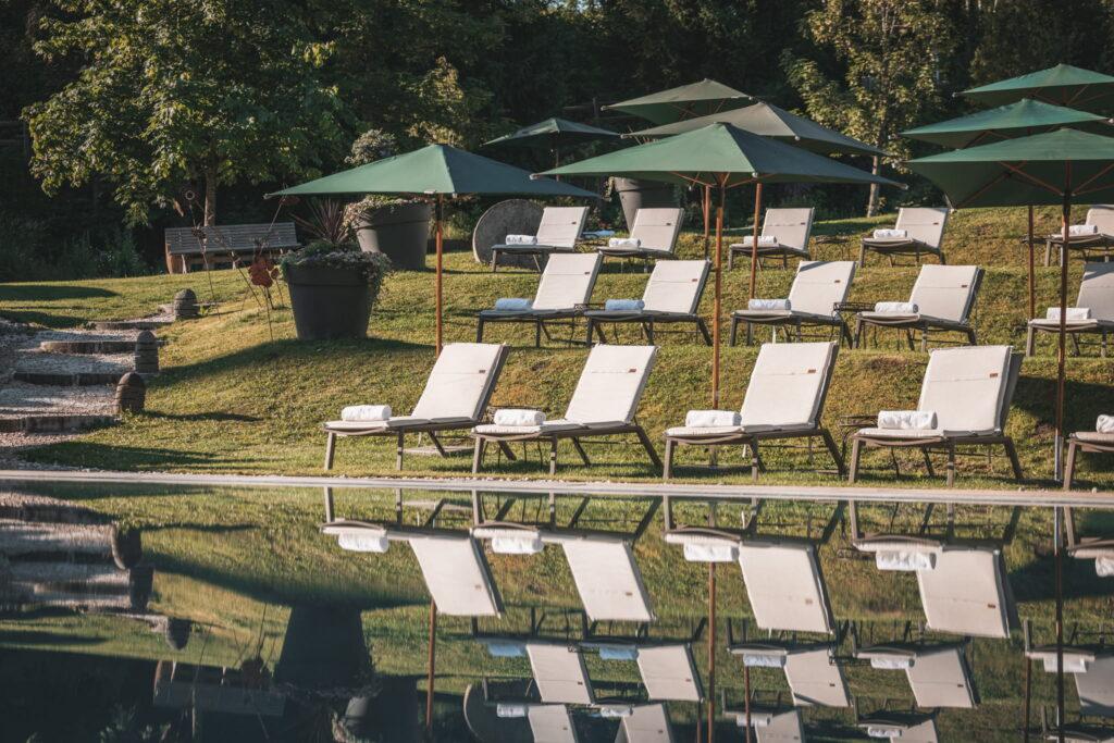 Rows of empty white deck chairs with green parasols stand on a well-kept lawn next to the tranquil outdoor pool of the sustainable Hotel Klosterbräu in Seefeld, radiating peace and relaxation under the sunny sky.