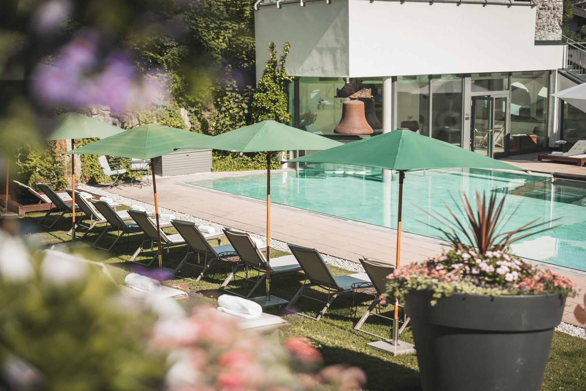 Rows of empty white deck chairs with green parasols stand on a well-kept lawn next to the tranquil outdoor pool of the sustainable Hotel Klosterbräu in Seefeld, radiating peace and relaxation under the sunny sky.