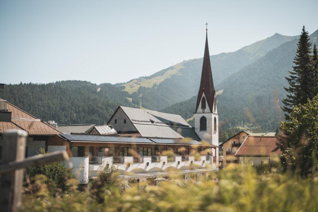 The picturesque village of Seefeld with a tall church tower against a backdrop of lush green mountains under a bright blue sky, radiating peace and serenity. The sustainable Hotel Klosterbräu can be seen in the foreground.