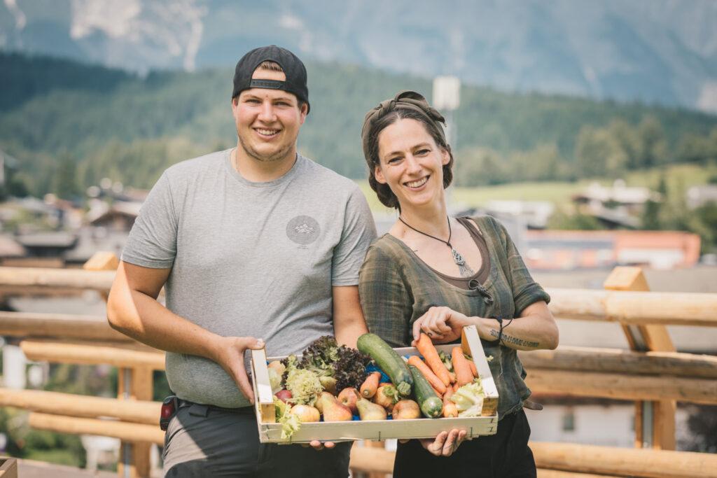 A smiling man and woman hold a basket of fresh vegetables, including carrots and zucchini, against the backdrop of mountains and rural surroundings in Seefeld, Austria. They are staying at the sustainable Hotel Klosterbräu.