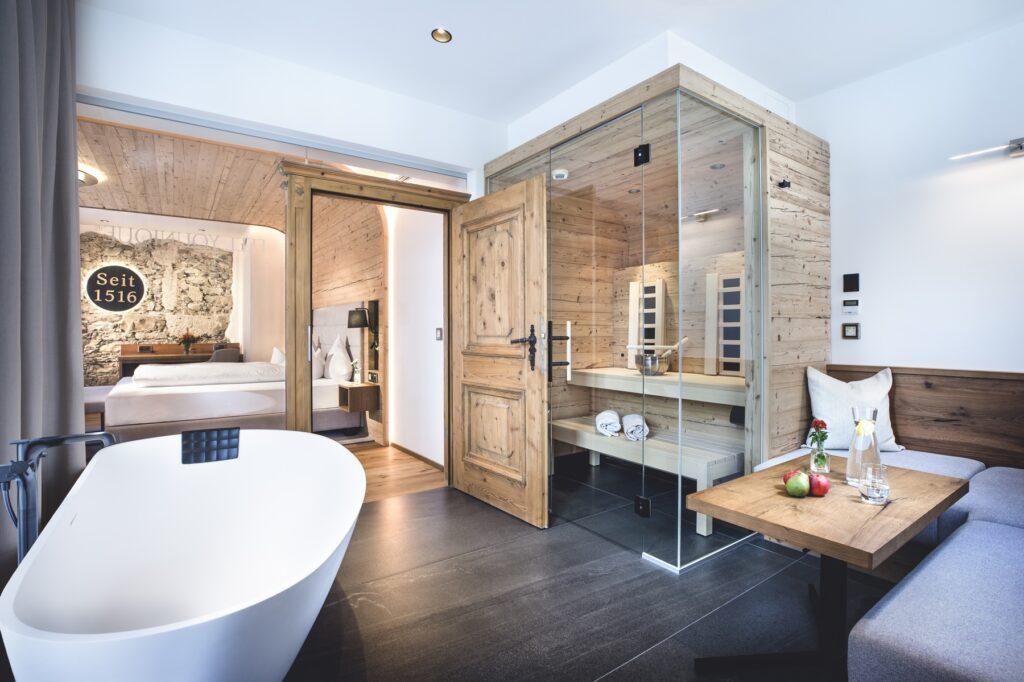 Modern hotel room in the sustainable wellness hotel Klosterbräu in Austria with a cozy atmosphere, freestanding bathtub, glass sauna, and spacious bed. Rustic wood decor provides warmth.