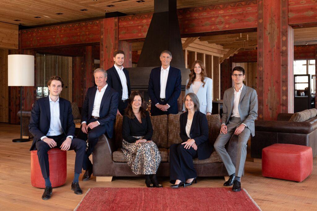 A group of eight professionally dressed family members of the Sanoner Family poses in a cozy, rustic room with wooden walls and decor. The mood is formal yet welcoming.