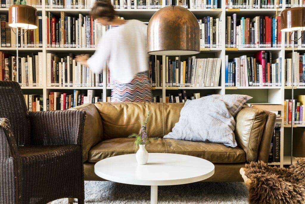 Gemütliche Bibliothek mit braunem Ledersofa, weißem runden Tisch und Bücherregalen im historischen und nachhaltigen Gut Sonnenhausen bei München. Eine verschwommene Person greift nach Büchern. Die Atmosphäre ist warm und einladend.