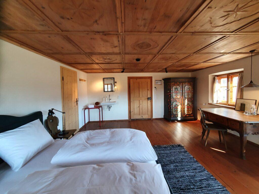 Gemütliches Zimmer aus Holz mit einem Bett, einem verzierten Kleiderschrank und einem rustikalen Schreibtisch im historischen Bauernhaus des Gut Sonnenhausen, einem nachhaltigen Hotel in der Nähe von München. Das warme Licht, das durch das Fenster fällt, unterstreicht die Vintage-Einrichtung und schafft eine ruhige Atmosphäre.