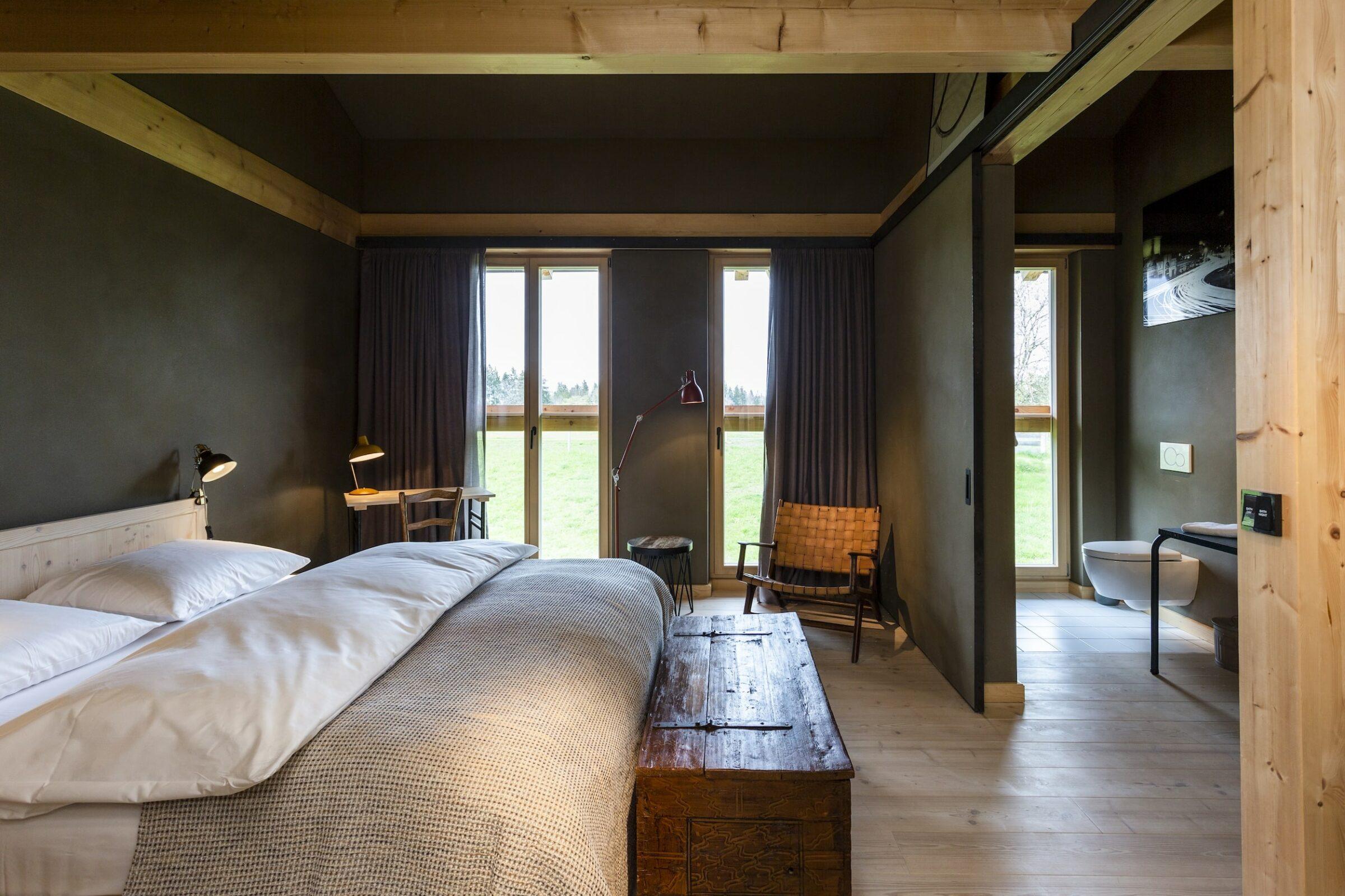 Gemütliches Schlafzimmer im Landhausstil mit großem Bett, Holzkommode und geflochtenem Stuhl im historischen Bauernhaus des Gut Sonnenhausen, einem nachhaltigen Hotel in der Nähe von München. Sonnenlicht fällt durch die Fenster mit dunklen Vorhängen und schafft eine warme, ruhige Atmosphäre.
