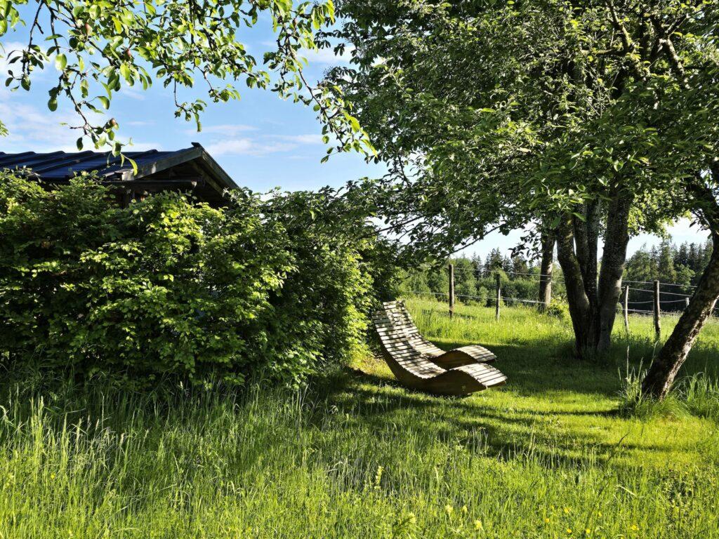 Ein Holzliegestuhl steht im Schatten von Laubbäumen, umgeben von saftigem grünem Gras und Büschen, mit einer ruhigen, sonnigen ländlichen Kulisse im Hintergrund. Erholung pur im nachhaltigen Hotel Gut Sonnenhausen