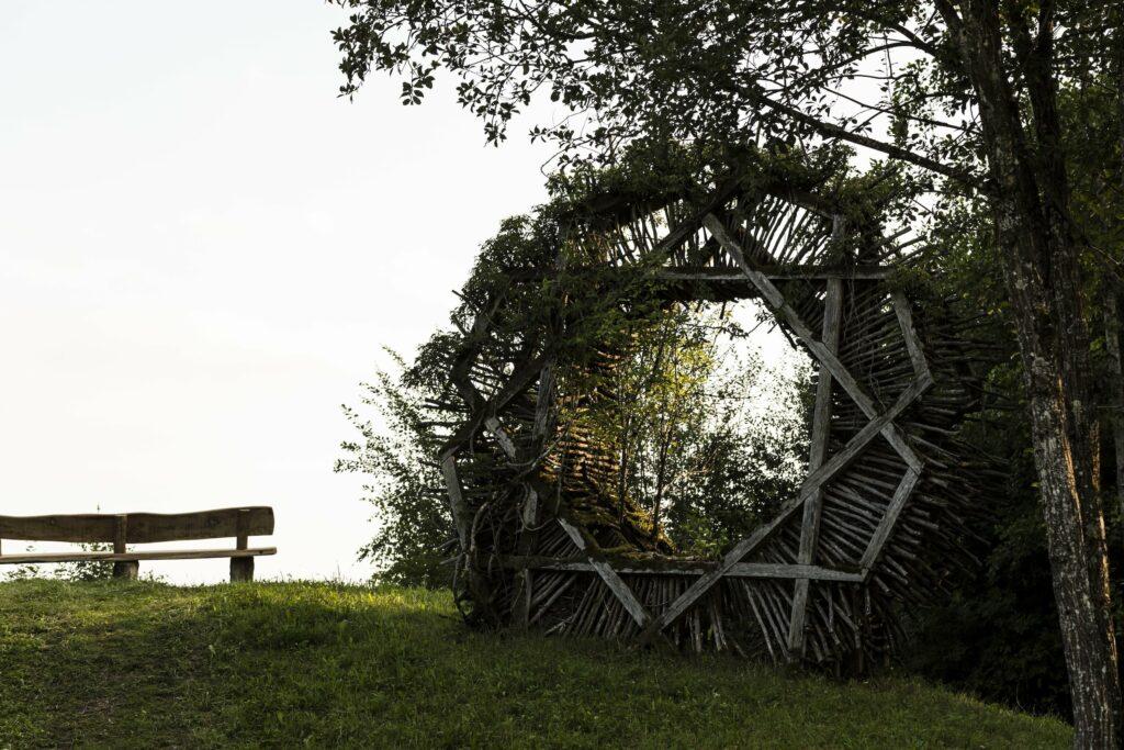 Auf einem grasbewachsenen Hügel steht eine rustikale Holzbank neben einem achteckigen Kunstwerk aus Holz, umgeben von viel Grün. Die Szene ist friedlich und ruhig – auf dem Gut Sonnenhausen in der Nähe von München.