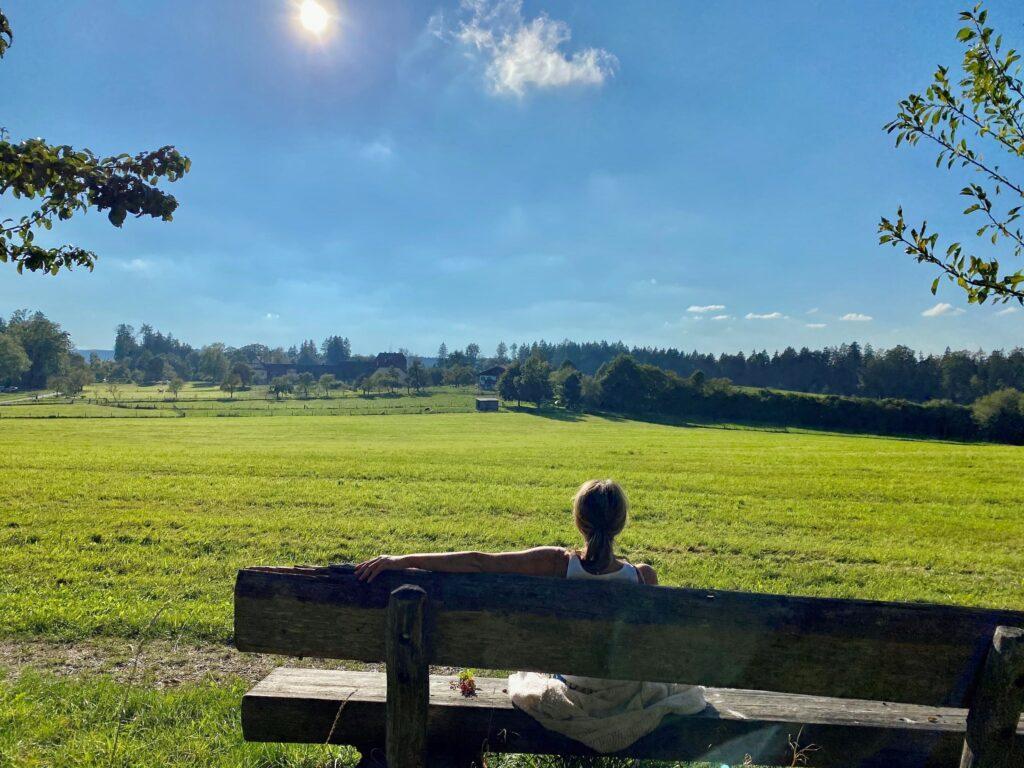 Eine Frau sitzt auf einer Holzbank mit Blick auf eine riesige grüne Wiese und das nachhaltige Hotel Gut Sonnenhausen unter einem strahlend blauen Himmel auf dem Sonnenhausener Höhenweg. Das Sonnenlicht sorgt für eine friedliche Atmosphäre und strahlt Ruhe und Entspannung aus.