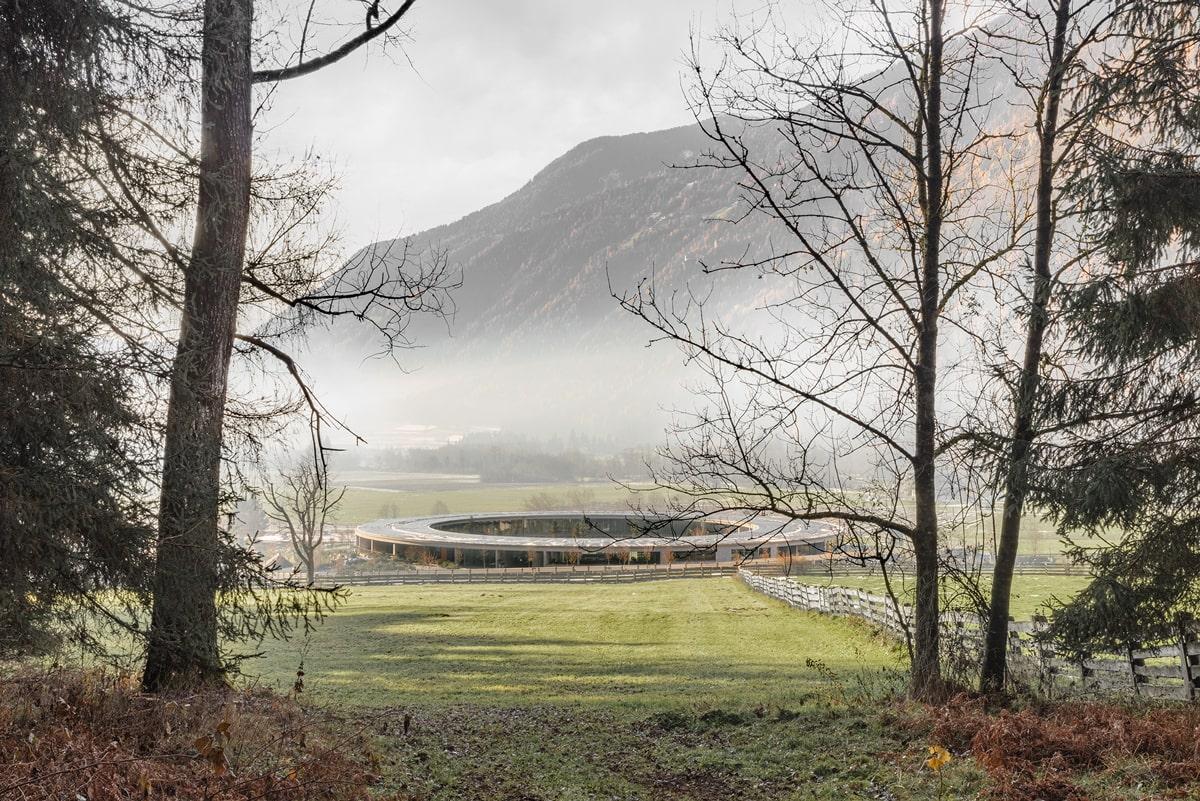 The circular building of the OLM Nature Escape sits in a misty valley of South Tyrol, surrounded by lush green fields and distant mountains. Bare trees frame the scene, conveying a serene, autumnal atmosphere.