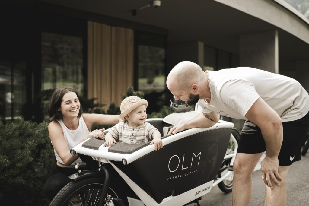 Young family getting ready for a ride on an electric cargo bike. On the side of the bike is OLM Nature Escape, a sustainable aparthotel in South Tyrol.