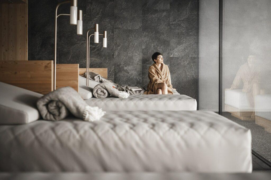 A person in a robe sits pensively on a modern bed in a simple, minimalist relaxation room at OLM Nature Escape. Soft morning light and a calm, contemplative mood.
