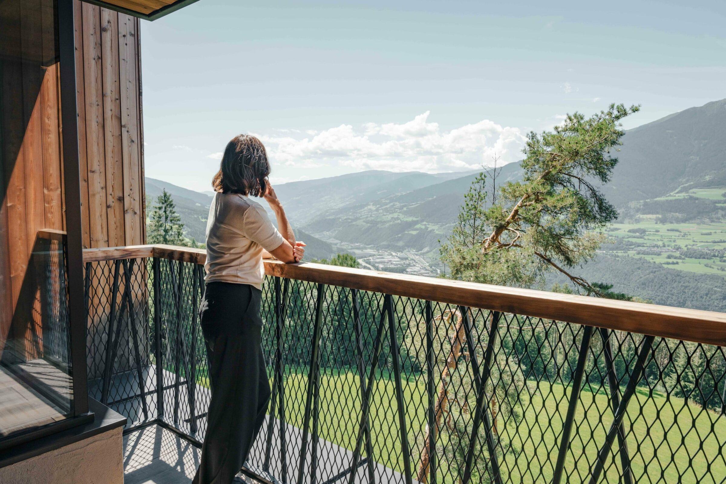 Eine Person in einem weißen Hemd und einer schwarzen Hose steht auf einem Holzbalkon des Hotels My Arbor, das nur Erwachsenen vorbehalten ist, und blickt unter einem strahlend blauen Himmel auf die malerische Aussicht auf die Berge und Täler.