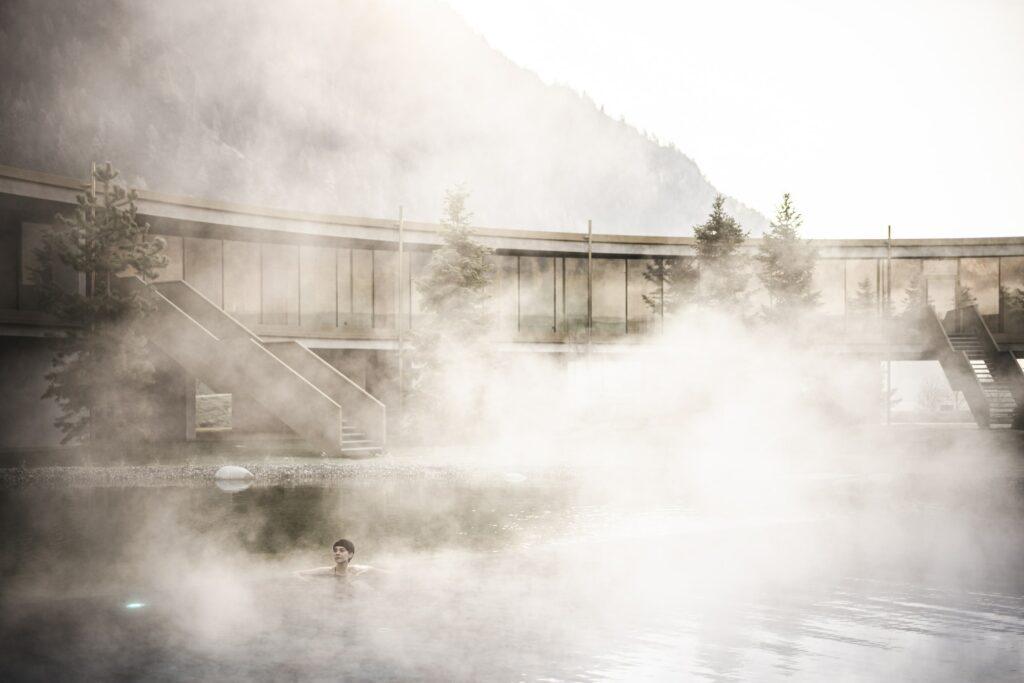 A serene scene with a person relaxing in the misty outdoor pool, surrounded by the modern architecture of the sustainable eco-apart-hotel OLM Nature Escape. Soft light and fog create a calm, ethereal atmosphere.