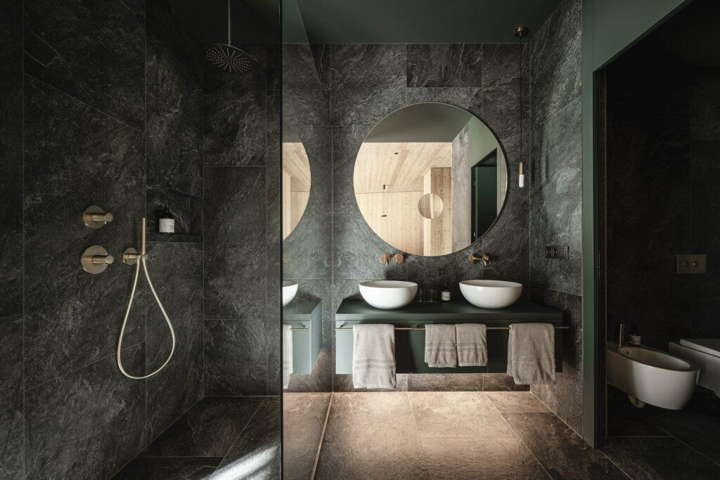 Luxurious bathroom of ApartSuite L in the sustainable apart-hotel OLM Nature Escape; with dark stone tiles, a rain shower, a double sink with round mirrors, white washbasins and towels that create a serene atmosphere.