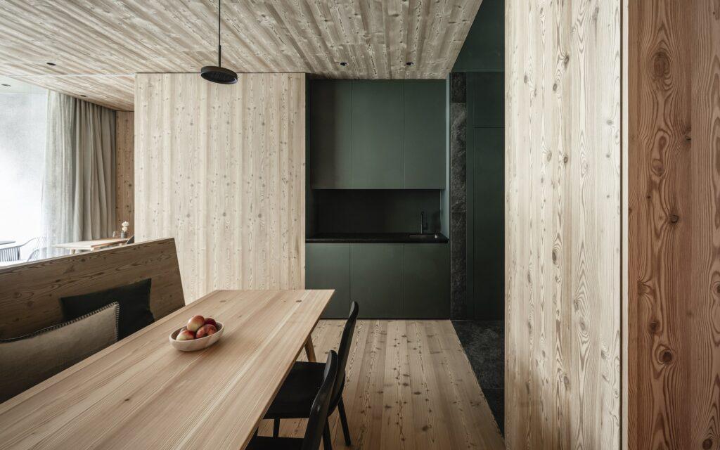 A modern, fully equipped kitchen with light-colored wooden paneling in the ApartSuites of the OLM Nature Escape eco-apart-hotel in South Tyrol. In front of it is a wooden dining table with a bowl of apples and black chairs. Dark green cabinets emphasize the minimalist style.