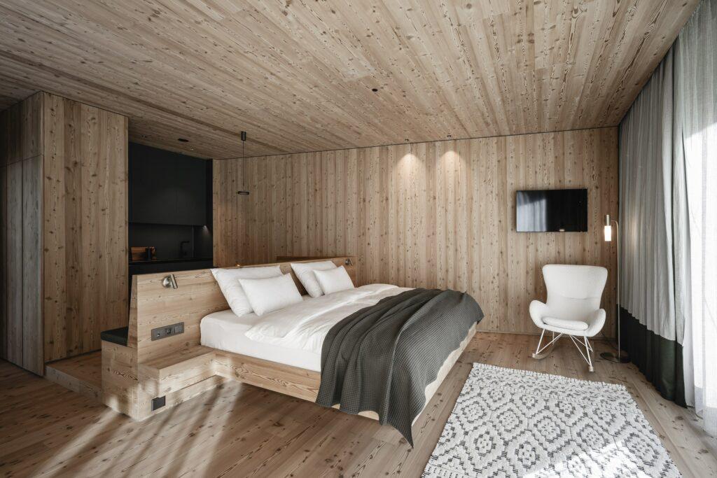 Stylish, modern bedroom in ApartSuite M of the eco-friendly apartment hotel OLM Nature Escape in South Tyrol; with wood-paneled walls and ceiling, minimalist bed, white chair and patterned rug. Soft lighting for a cozy ambiance.