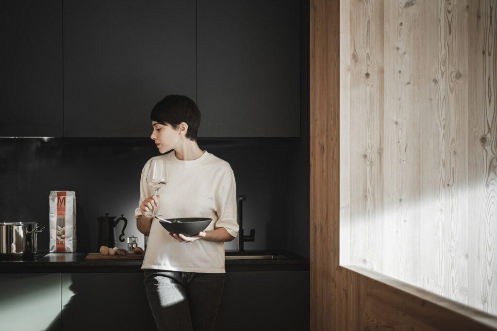 A woman is seen holding a wine glass and a bowl in the modern kitchenette of an apart-suite at the OLM Nature Escape eco-apart-hotel in South Tyrol. She is standing near a stove. Soft light from a wooden window creates a calm, cozy atmosphere.