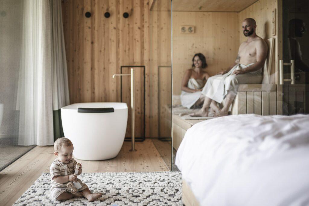 Cozy wooden ApartSuite for families with bathtub and sauna in the sustainable hotel OLM Nature Escape. A baby plays on a rug while two adults, smiling and wrapped in towels, relax in the sauna. Warm and calm atmosphere.