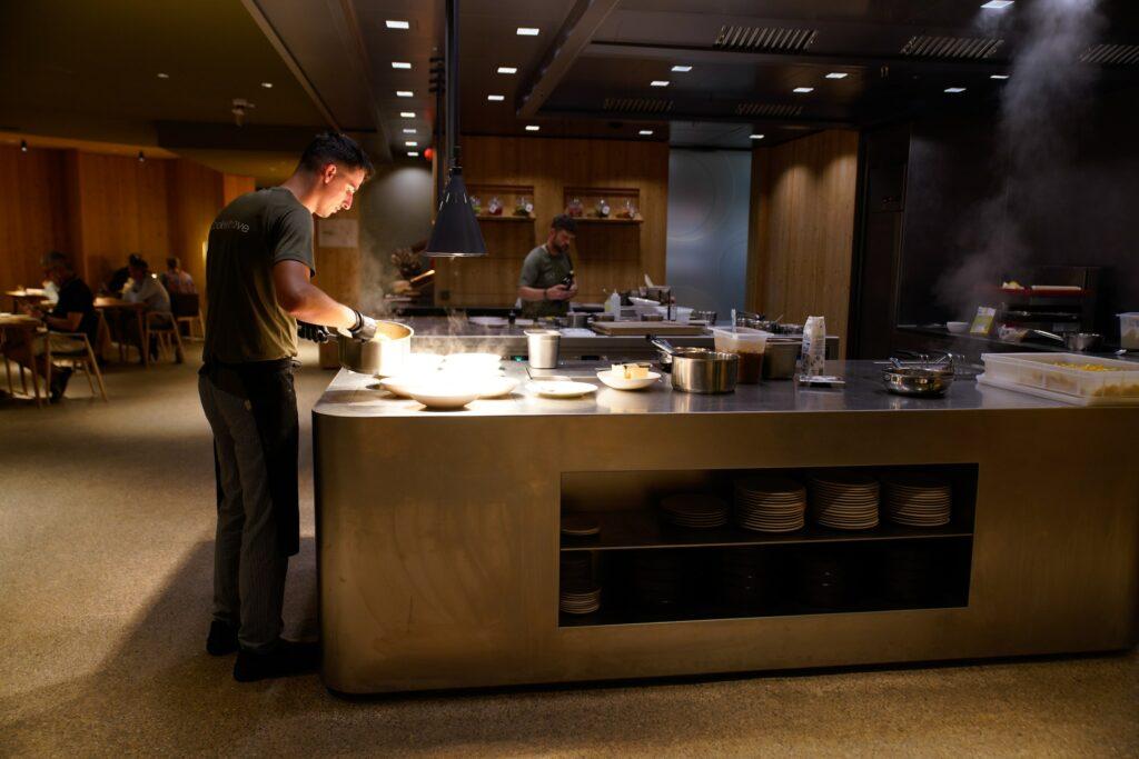 A chef prepares food at the modern open kitchen counter in the sustainable restaurant PRENN° of the OLM Nature Escape. Warm lighting, guests dine in the background. The scene is cozy and inviting.
