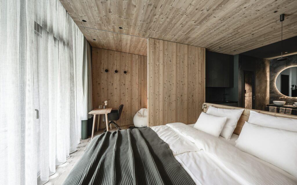 Modern and sustainable suite with a separate bathroom in the OLM Nature Escape with wood-paneled walls and ceiling, furnished with a cozy bed with crisp white linens. A small desk and chair are set near sheer curtains, creating a calm atmosphere.