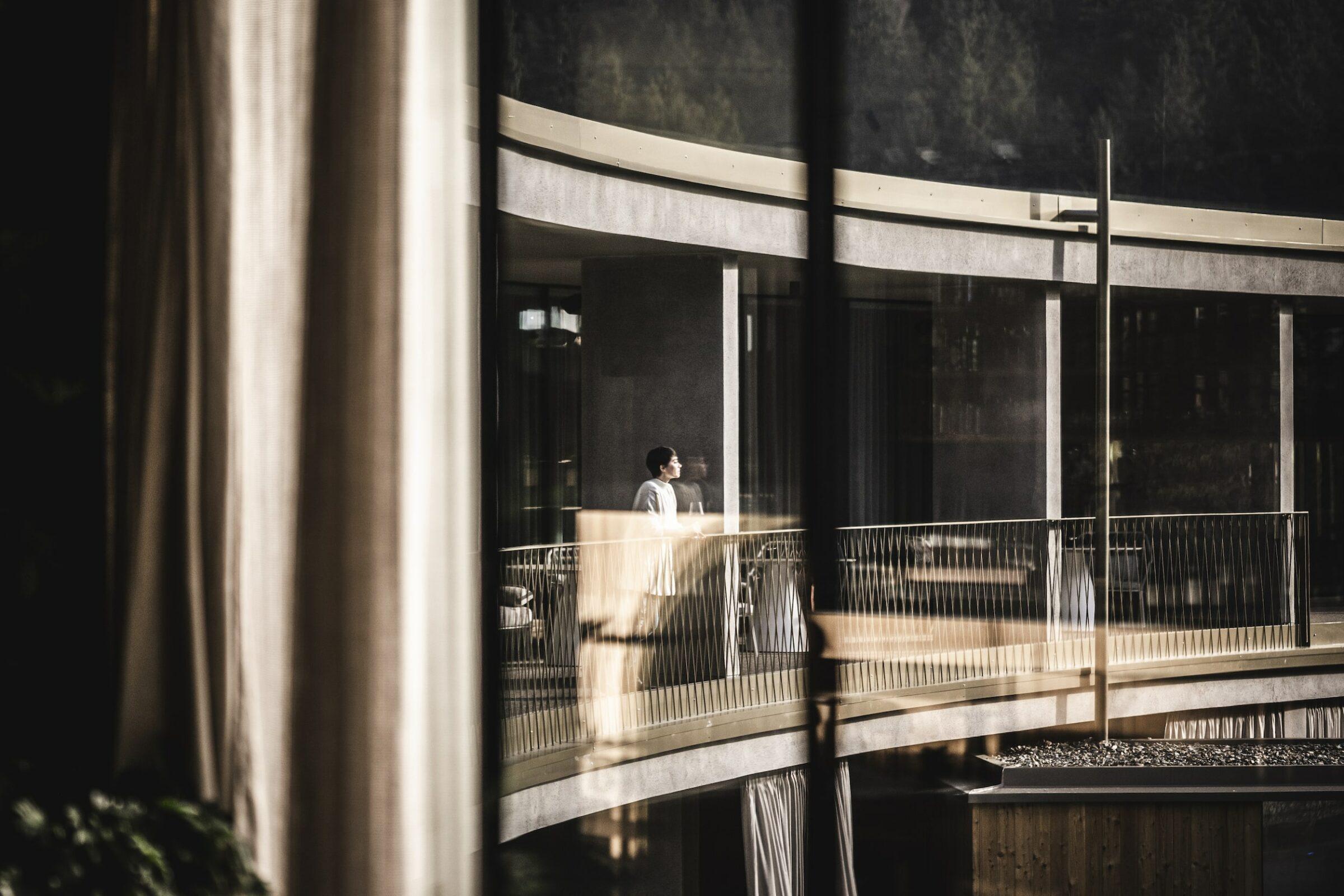 A person stands on the curved balcony of their ApartSuite in the sustainable hotel OLM Nature Escape and is observed through a window with reflections. Soft light and shadows create a calm, contemplative atmosphere.