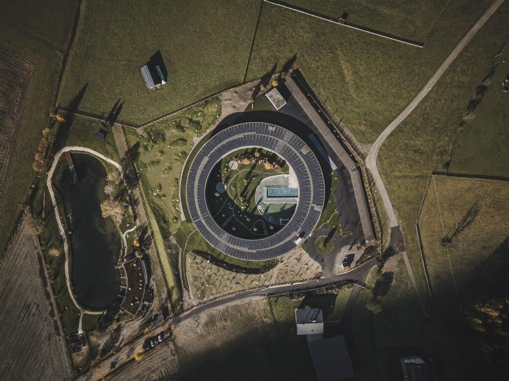 Aerial view of the round building of the OLM Nature Escape eco-aparthotel, which has solar panels and is located in the middle of a lush green landscape. There is a pond with trees nearby, creating a harmonious backdrop.