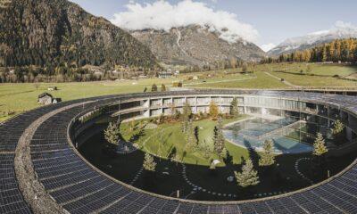 Das runde Gebäude des Eco Aparthotel OLM Nature Escape mit Solarmodulen auf dem Dach umschließt einen Innenhof mit Bäumen und einem Pool. Es liegt vor einer Kulisse aus grünen Feldern, Bergen und einem bewölkten Himmel und vermittelt ein Gefühl der Harmonie mit der Natur.