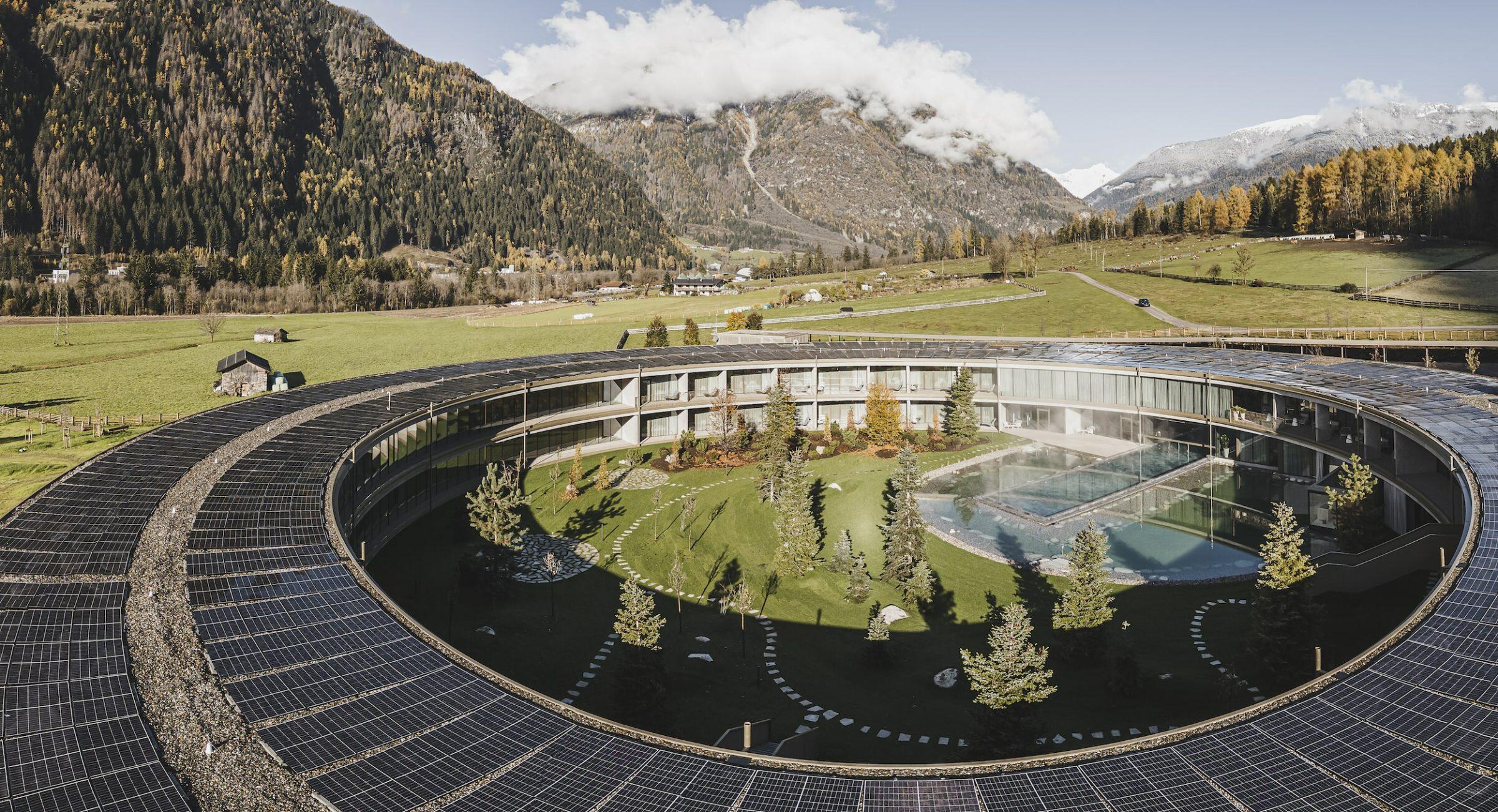 The round building of the Eco Aparthotel OLM Nature Escape, with solar panels on the roof, encloses a courtyard with trees and a pool. It is set against a backdrop of green fields, mountains and a cloudy sky, conveying a sense of harmony with nature.