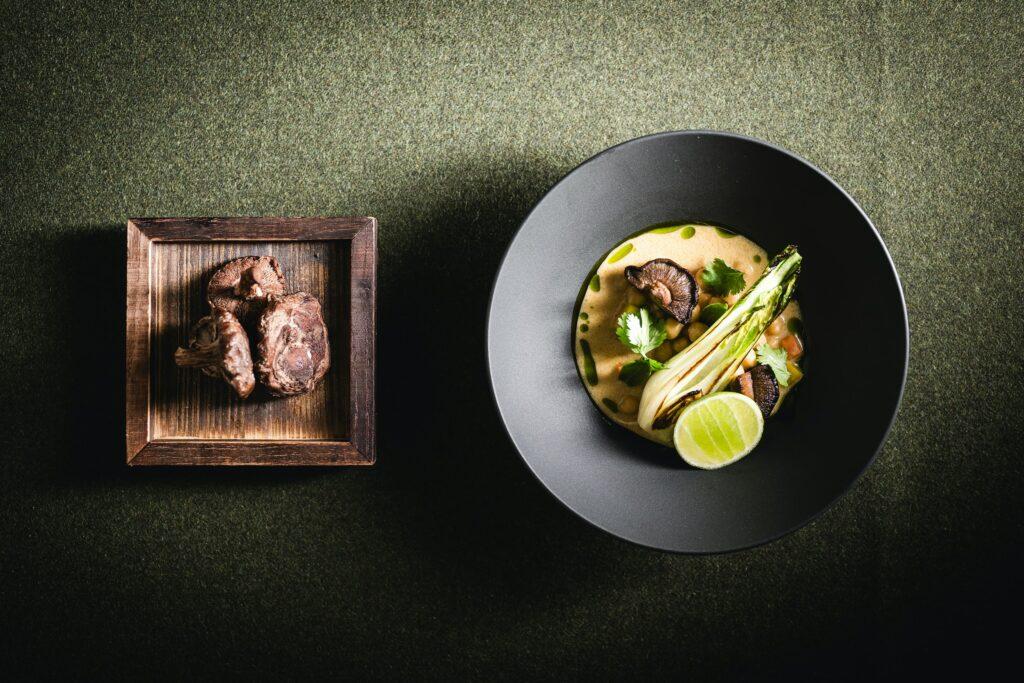 A black bowl of creamy hummus, garnished with mushrooms, pak choi, coriander and a slice of lime, is placed on a green area next to a wooden box of shitake mushrooms.