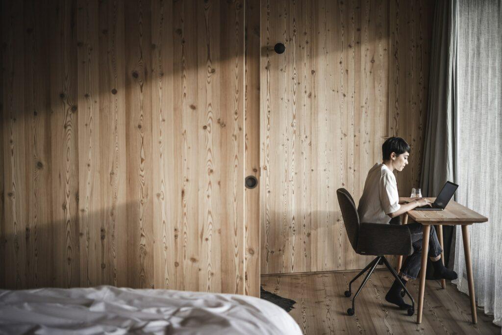 A person sits at a wooden table and works on a laptop in a minimalist ApartSuite with wooden walls at the sustainable hotel OLM Nature Escape. Soft light falls through a window, creating a calm atmosphere.
