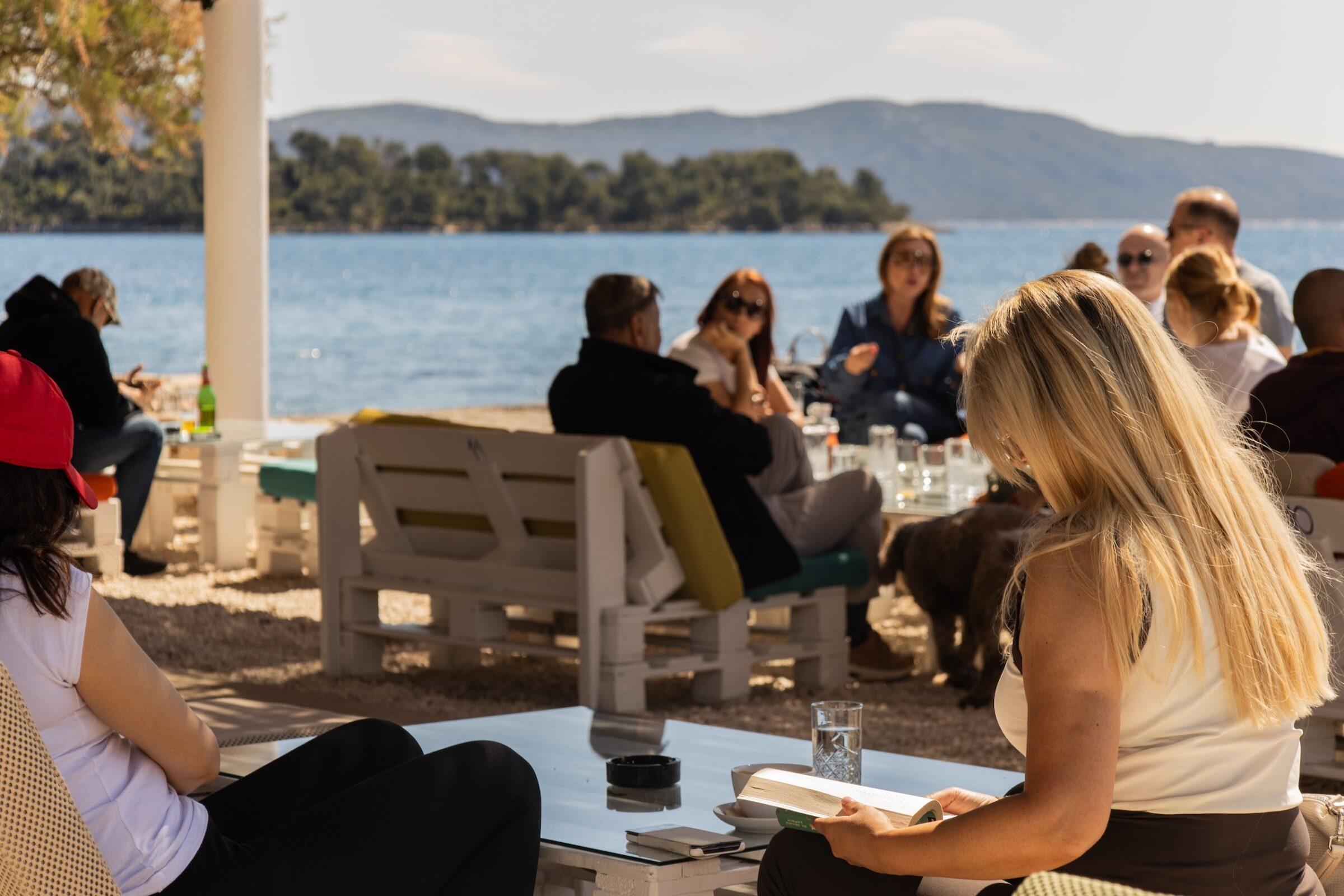 Mehrere Personen sitzen in einer Outdoor-Lounge des Hotels Korinjak in Kroatien an weißen Holzmöbeln mit Blick auf das Meer, eine Frau im Vordergrund liest ein Buch.