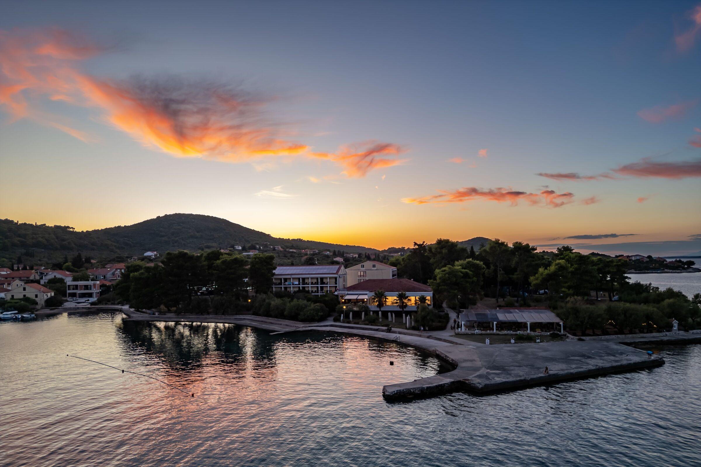 Hotel Korinjak an der kroatischen Küste bei Sonnenuntergang mit ruhigem Wasser, bewaldeten Hügeln und Gebäuden am Ufer