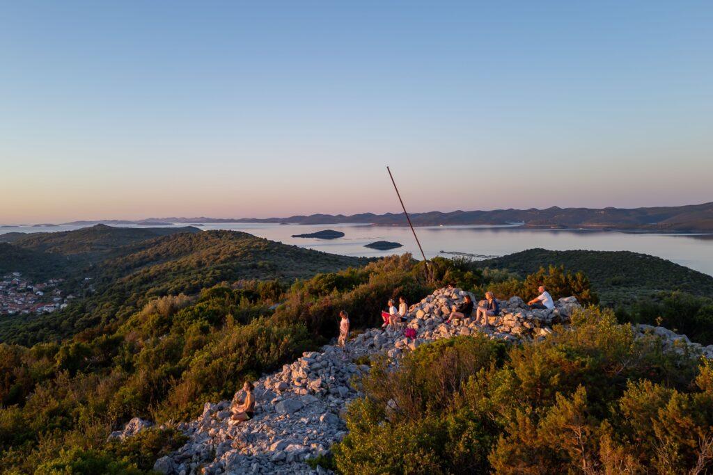 Gruppe von Menschen sitzt auf felsigem Hügel mit Blick auf bewaldete Landschaft und Meer bei Sonnenaufgang in Kroatien