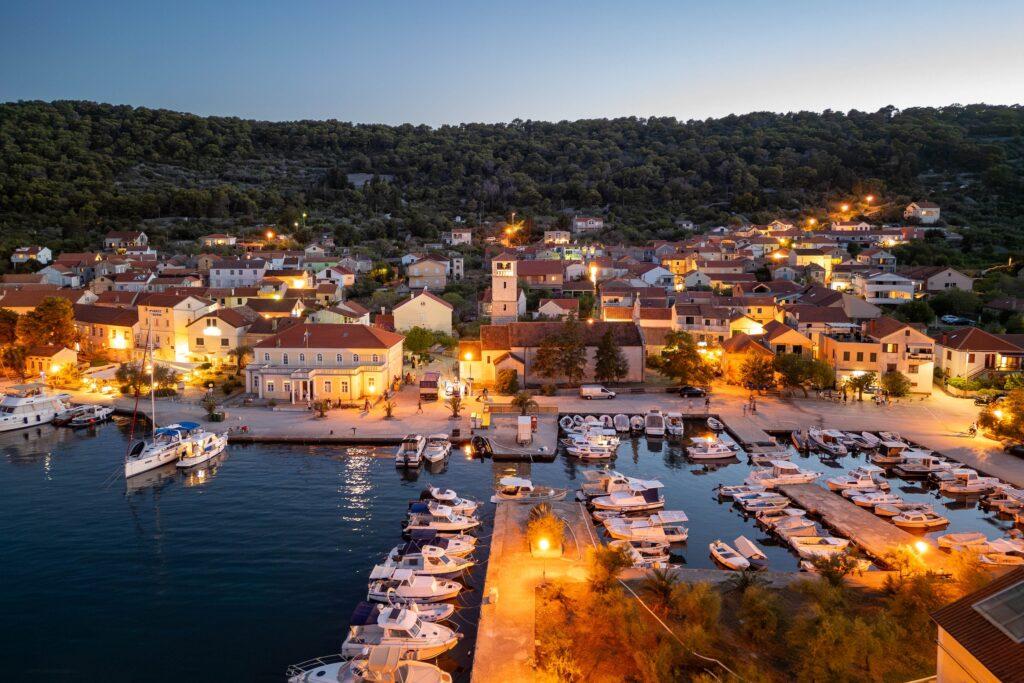 Nachtaufnahme des Hafens von Veli Iz in Kroatien mit beleuchteten Booten, Gebäuden und bewaldetem Hügel im Hintergrund
