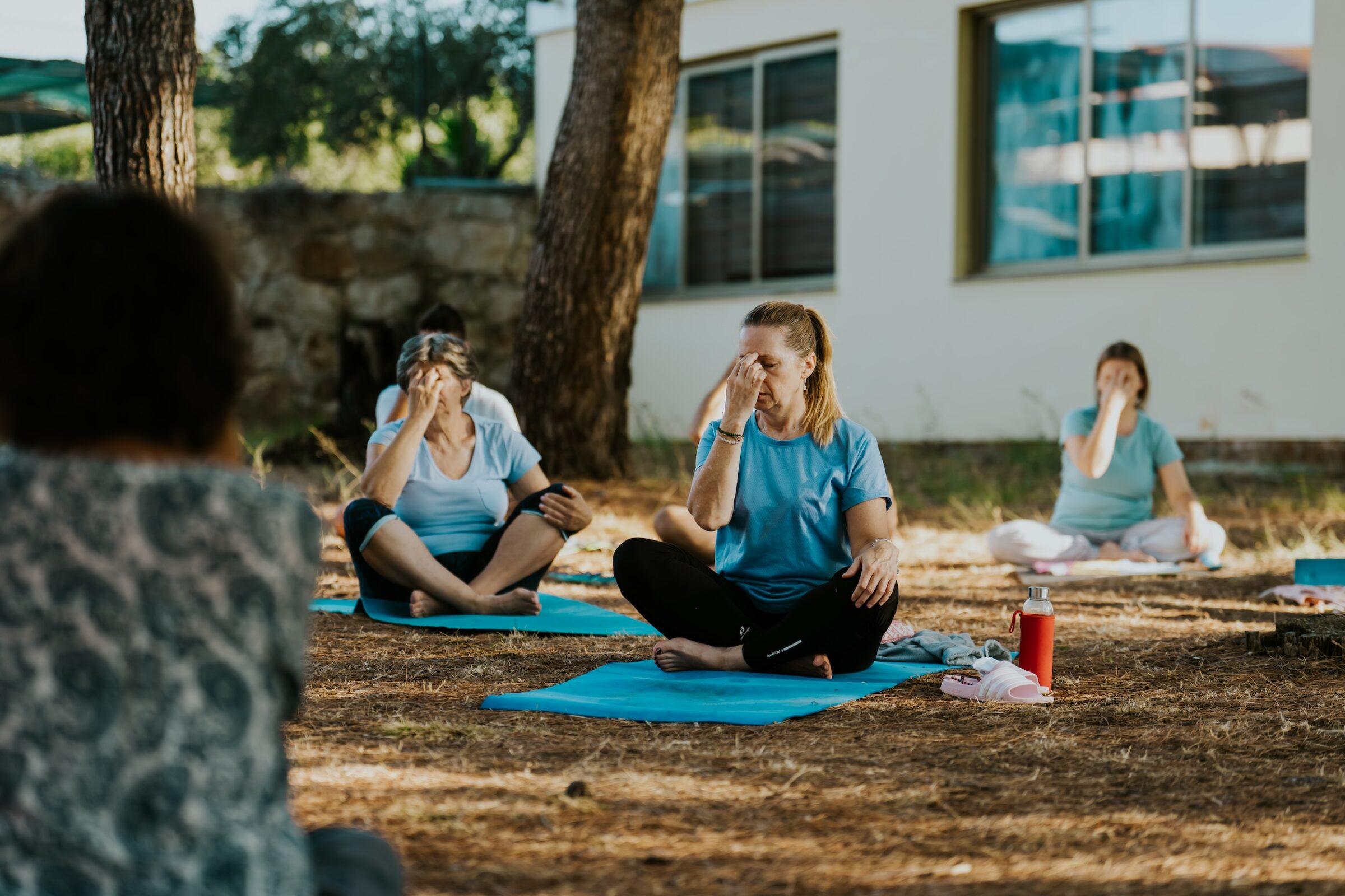 Mehrere Personen sitzen auf Yogamatten im Freien vor einem Gebäude und praktizieren Atemübungen, wobei sie jeweils eine Nasenöffnung mit der Hand verschließen