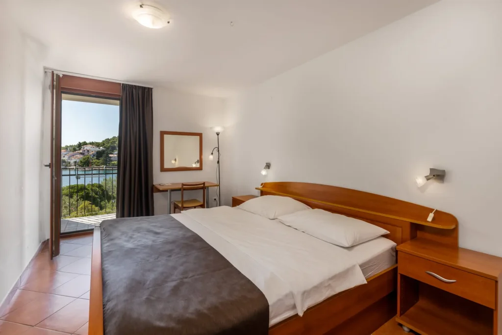 Doppelzimmer im Hotel Korinjak mit Doppelbett, Nachttischen, Schreibtisch, Stuhl, Stehlampe und Balkon mit Blick auf das Meer und umliegende Häuser