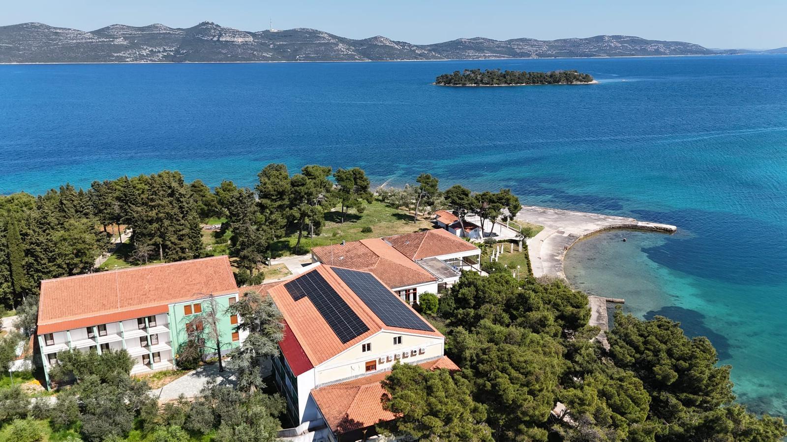 Aerial view of a coastal complex with the red roofs and solar panels of the vegetarian Korinjak hotel, surrounded by lush trees and a bright blue sea. Small island visible in the distance.