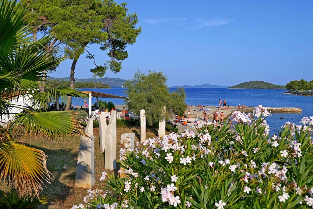 Küstenszene in Kroatien mit leuchtend blauem Meer, Menschen, die sich an einem Sandstrand unter einem klaren Himmel entspannen, eingerahmt von üppigem Grün und rosa Blumen im Vordergrund. Nahe des nachhaltigen Hotel Korinjak.