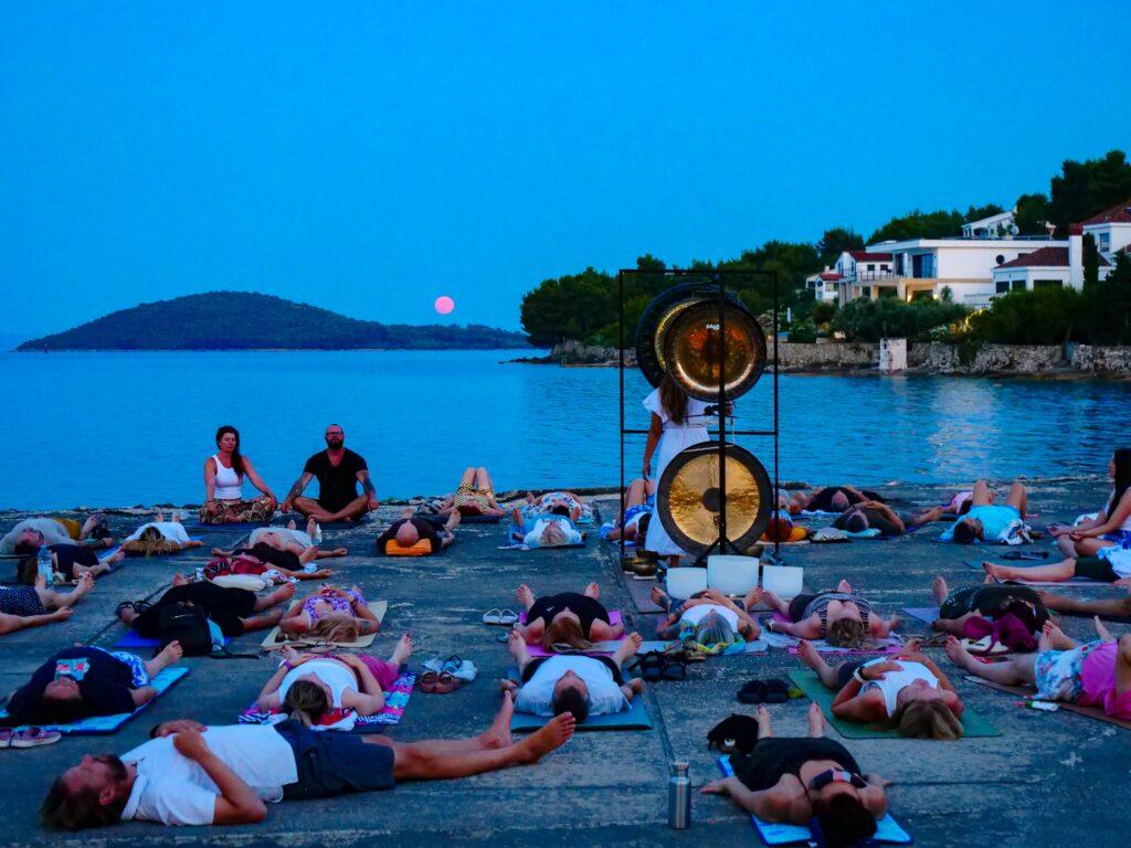 Gruppe von Menschen liegt auf Yogamatten am Meer bei Hotel Korinjak in Kroatien während eines Gong-Klangbads im Vollmondlicht mit zwei großen Gongs im Vordergrund