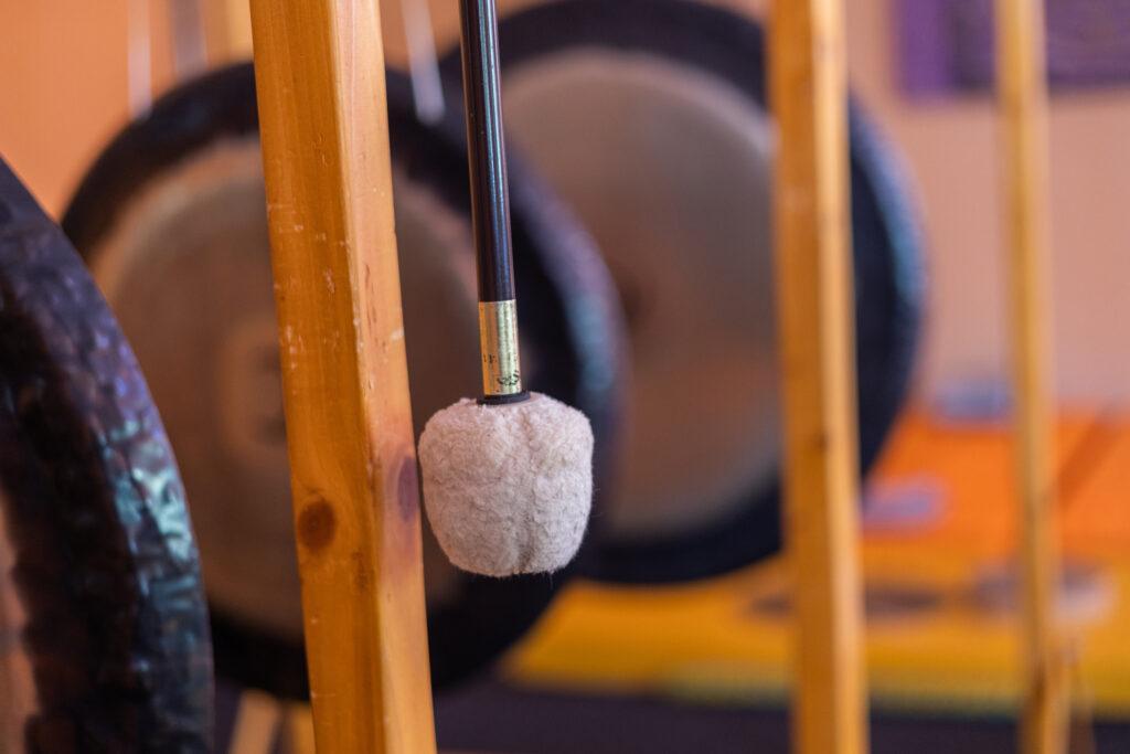 Nahaufnahme eines Gong-Schlägels mit einem flauschigen, runden Kopf, der auf einem Holzrahmen ruht. Verschwommene Gongs im Hintergrund erzeugen eine ruhige, meditative Atmosphäre. Willkommen im ganzheitlichen Hotel Korinjak in Kroatien.