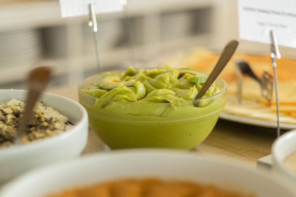 Eine Glasschüssel mit einer glatten, cremigen grünen Avocadocreme und einem Löffel darin. Die umliegenden Gerichte auf einem Holztisch bilden ein warmes, einladendes Buffet im vegetarischen Hotel Korinjak in Kroatien