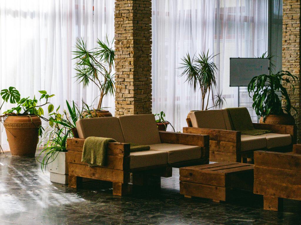 The spacious lobby of the sustainable Hotel Korinjak features stone columns, white curtains, wooden sofas and potted plants, creating a calm, natural atmosphere with warm, earthy tones.