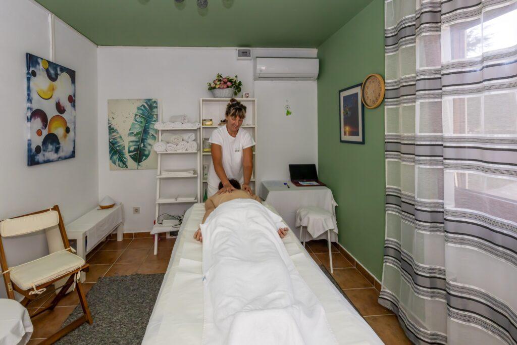 Massageraum im Hotel Korinjak in Kroatien mit einer liegenden Person auf der Massageliege und einer Masseurin, die eine Rückenmassage durchführt, umgeben von grünen Wänden, einem Fenster mit gestreiften Vorhängen und einfacher Einrichtung