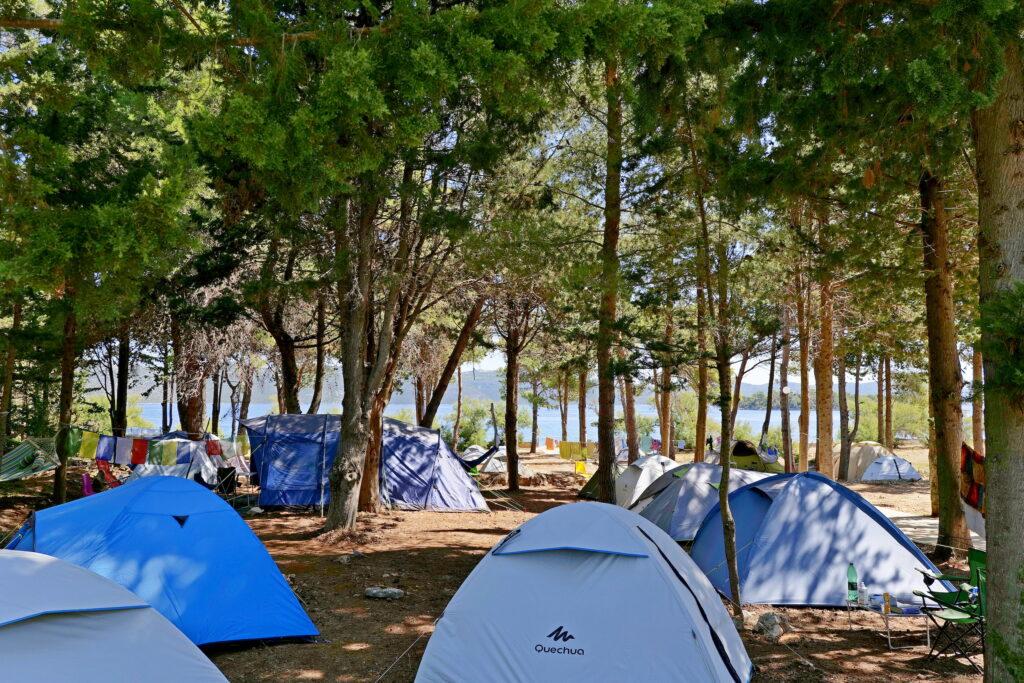 Der ruhige Waldcampingplatz des nachhaltigen Hotel Korinjak in Kroatien mit mehreren blauen und grauen Zelten. Das Sonnenlicht dringt durch die Bäume, und im Hintergrund ist ein See zu sehen, der Ruhe ausstrahlt.