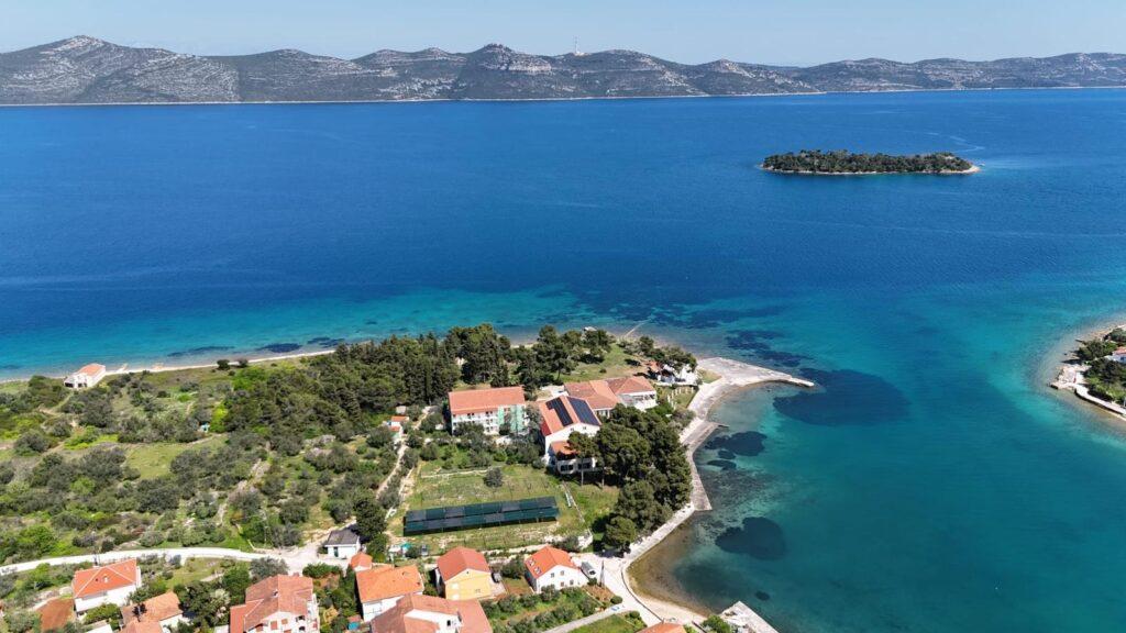 Luftaufnahme einer Küstenlandschaft auf der kroatischen Insel Iz mit türkisfarbenem Wasser, üppigem Grün und verstreuten Häusern mit roten Dächern, darunter das vegetarische Hotel Korinjak. Eine Insel und ferne Berge sind zu sehen.