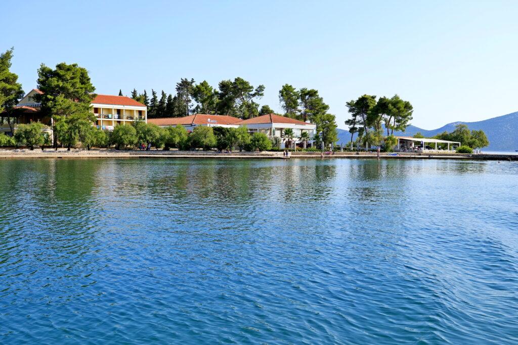 Das ruhige, nachhaltige Hotel Korinjak am Meer, umgeben von üppigen Bäumen, mit ruhigem, blauem Wasser im Vordergrund und fernen Bergen unter einem klaren blauen Himmel.