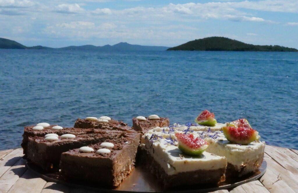 Scheiben von dunklem Schokoladenkuchen und hellem Kuchen mit Feigen (raw food) auf einem Steg vor dem Hotel Korinjak. Im Hintergrund ist das ruhige kroatische Meer mit Hügeln unter einem blauen Himmel zu sehen.