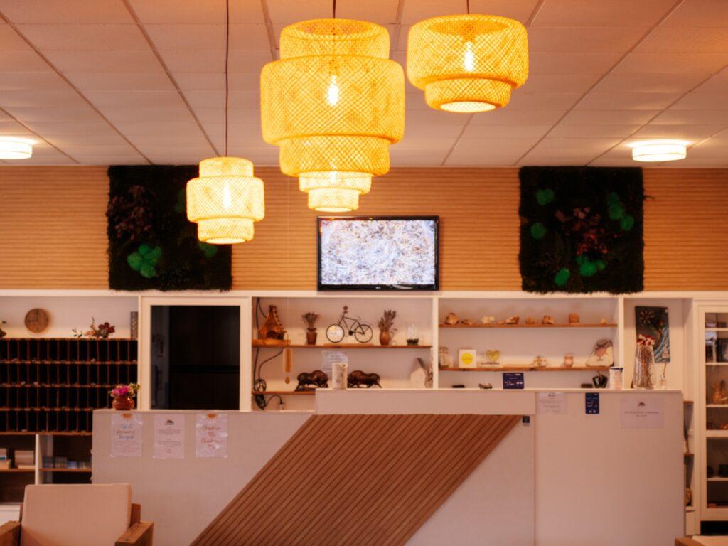 Gemütliche Lobby im vegetarischen, kroatischen Hotel Korinjak mit warmer Beleuchtung durch geflochtene Hängelampen. Ein Empfangstresen mit Holzmuster befindet sich vor Regalen mit Dekoration. Darüber ist ein Fernseher angebracht, und die Wände sind mit grünen Pflanzenakzenten geschmückt, die eine einladende Atmosphäre schaffen.